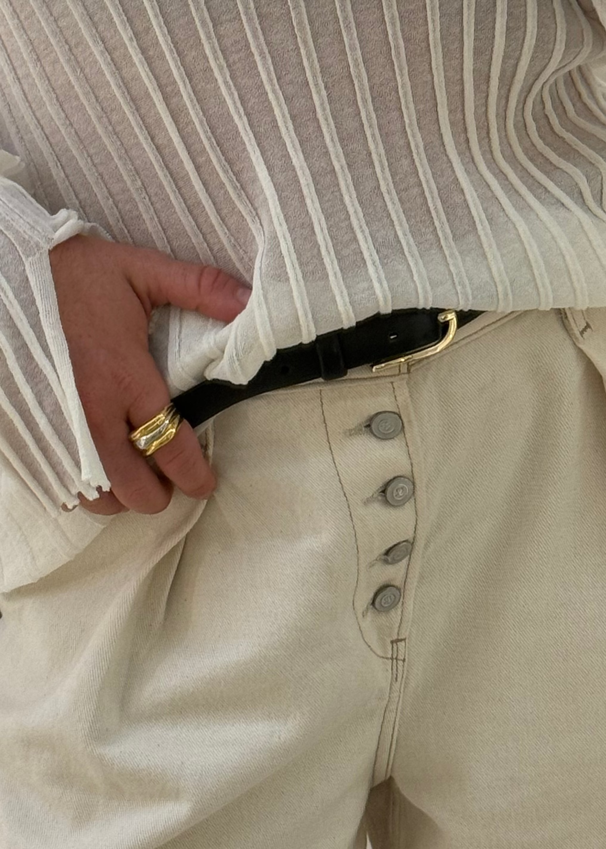 Close-up of a person wearing a striped shirt and beige pants with a gold ring on a neutral background